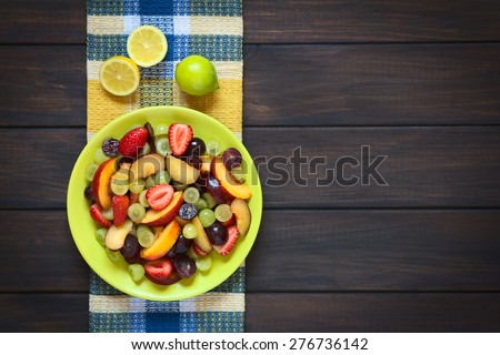 Fresh fruit salad made of grape, strawberry, plum and nectarine served on plate with lemon above, photographed overhead on dark wood with natural light