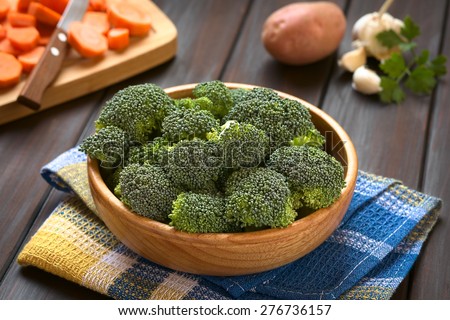 Fresh raw broccoli florets in wooden bowl, carrot slices, potato, garlic, parsley in back, photographed on dark wood with natural light (Selective Focus, Focus one third into the broccoli)