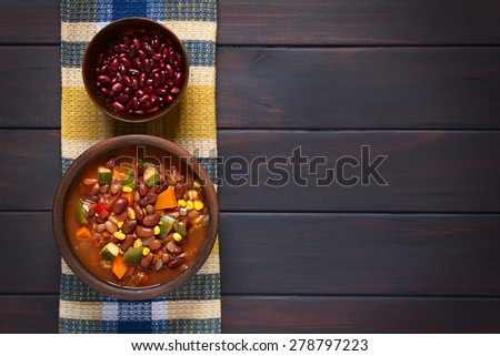 Vegetarian chili dish made of kidney bean, carrot, zucchini, bell pepper, sweet corn, tomato, onion, garlic in bowl, raw kidney beans above, photographed overhead on dark wood with natural light