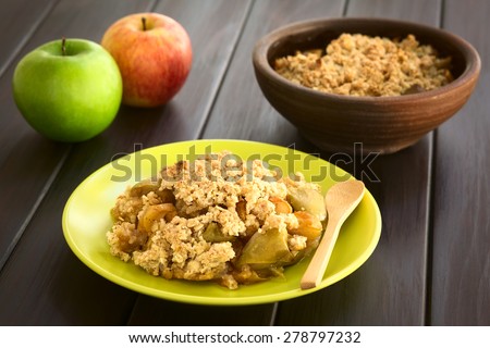 Freshly baked apple crumble or crisp served on plate with wooden spoon, fresh apples in the back, photographed on dark wood with natural light (Selective Focus, Focus one third into the crumble)