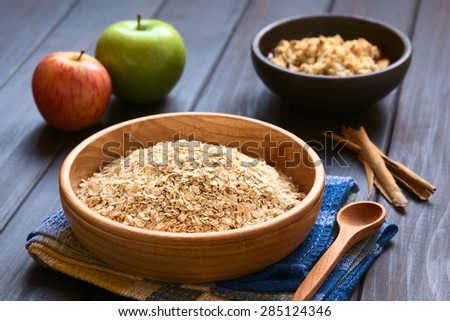 Raw rolled oats in wooden bowl with apples, cinnamon sticks and a bowl of fruit crumble in the back, photographed on dark wood with natural light (Selective Focus, Focus one third into the oats)