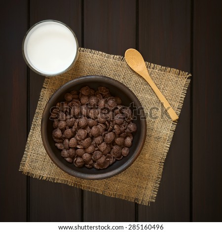 Crispy chocolate corn flakes breakfast cereal in rustic bowl with a glass of milk and wooden spoon on the side, photographed overhead on dark wood with natural light