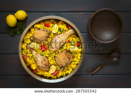 Chicken paella, a traditional Valencian (Spanish) rice dish made of rice, chicken, peas and capsicum and served with lemon in  pot, photographed overhead on dark wood with natural light