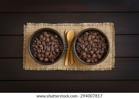 Crispy chocolate corn flakes breakfast cereal in rustic bowls with small wooden spoons, photographed overhead on dark wood with natural light
