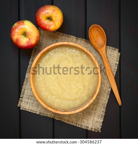 Homemade apple sauce in wooden bowl with wooden spoon and fresh apples on the side, photographed overhead on dark wood with natural light