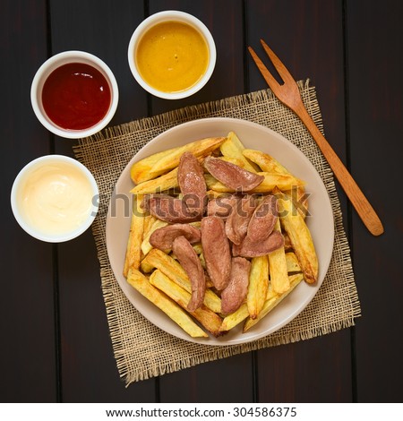 Salchipapas made of French fries and fried sausage, traditional fast food in South America, mayonnaise, ketchup, mustard and fork on the side, photographed overhead on dark wood with natural light