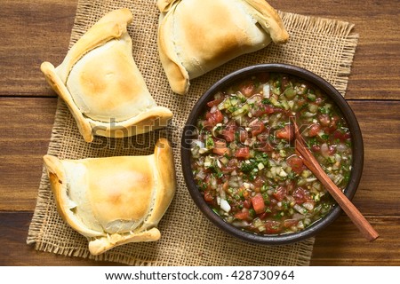 Chilean Pebre sauce, a traditional condiment made of tomato, onion, garlic, spicy aji pepper and coriander with empanadas on the side, photographed overhead on dark wood with natural light