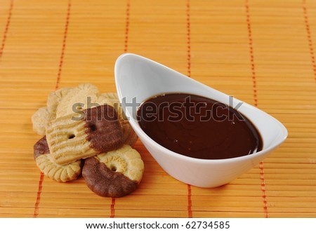 Butter cookies dipped in milk chocolate icing with chocolate dip in white bowl on orange-colored table mat (Selective Focus, Focus on front rim of bowl and part of the icing and the front cookie)