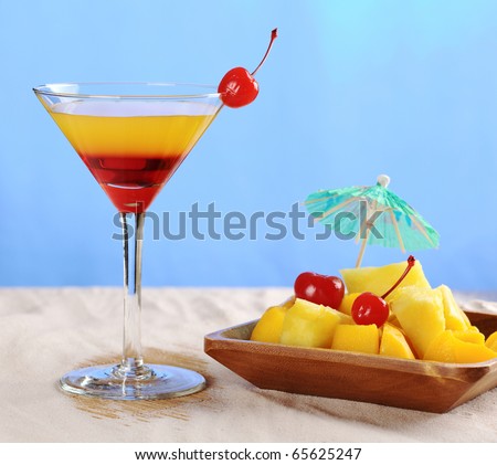 Yellow-red colored cocktail with cherry and tropical fruits (pineapple, mango) in wooden bowl on white sand (Selective Focus, Focus on the cherry on the glass, parts of the glass rim and the fruits)