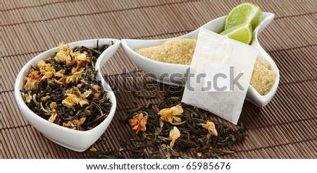 Teabag with an empty label, green tea with jasmine flowers, and brown sugar with lemon pieces in bowl on brown table mat (Selective Focus, Focus on the teabag)