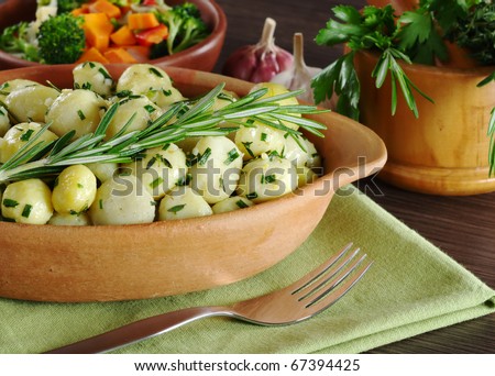 Small potatoes with herbs (parsley, thyme, rosemary) with a fork; with fried vegetables, garlic and fresh herbs in a wooden mortar in the background (Selective Focus, Focus on the rosemary branch)
