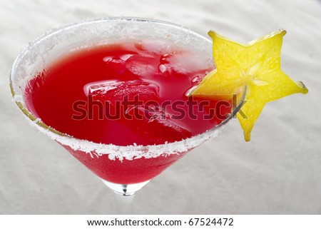 Red cocktail with ice cubes and garnished with carambola slice and coconut flakes on the rim, photographed on white sand (Selective Focus, Focus on the carambola slice)