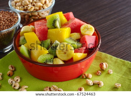 Fresh fruit salad made of banana, kiwi, watermelon and mango pieces in orange bowl with cereals (puffed wheat and quinoa) (Selective Focus, Focus on the front of the bowl and the fruits in the front)