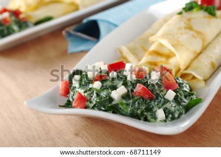 Spinach-tomato-cheese sauce with pancakes on a long plate with cutlery wrapped in blue napkin on wood (Selective Focus, Focus on the front of the sauce)