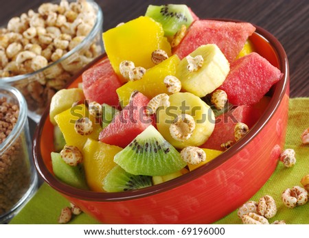 Fresh fruit salad made of banana, kiwi, watermelon and mango pieces in orange bowl with cereals (puffed wheat and chocolate quinoa) (Selective Focus, Focus on the front of the bowl and the fruits)