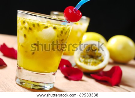 Passion fruit juice with a drinking straw and a maraschino cherry as well as passion fruit and rose petals in the background on wooden board (Selective Focus, Focus on the maraschino cherry)
