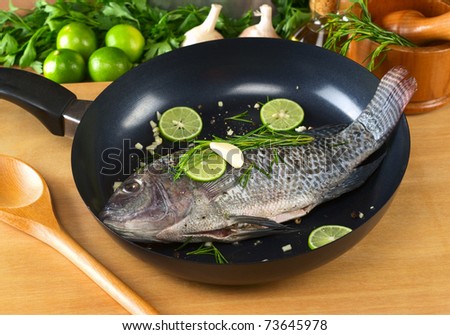 Raw tilapia with condiments (pepper corns, lime slices, garlic and rosemary) in frying pan with a wooden stirring spoon (Selective Focus, Focus on the head and belly of the fish)