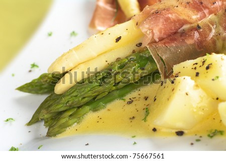 White and green asparagus wrapped in thin ham slices with Hollandaise sauce on top garnished with black pepper and parsley with cooked potato in front (Selective Focus, Focus on the front)