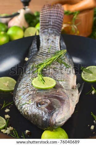 Raw tilapia with condiments (pepper corns, lime slices, garlic and rosemary) in frying pan (Selective Focus, Focus on the eye)