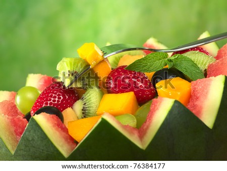 Fresh fruit salad (strawberry, kiwi, mango, grape) in melon bowl with kiwi and mango on fork and mint leaf as garnish (Selective Focus, Focus on the fruit on the fork and the mint leaf)