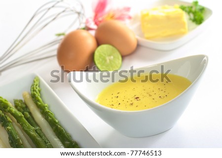 Hollandaise sauce with black pepper, asparagus on the side and the ingredients (butter, egg, lemon) of the sauce in the back (Selective Focus, Focus in the middle of the bowl)
