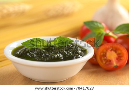 Fresh pesto made of basil, garlic and olive oil in a bowl and garnished with basil leaf with spaghetti, cherry tomatoes, basil and garlic in back (Selective Focus, Focus on the basil leaf in the bowl)