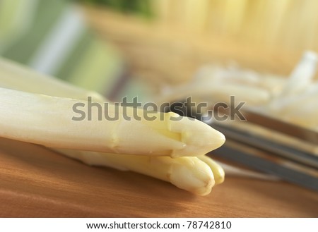 Two white asparagus with peeler and asparagus peel in the back on wooden board (Very Shallow Depth of Field, Focus on the two asparagus heads)