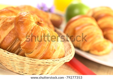 Fresh croissants in bread basket with a red knife beside, and a plate with croissants, green apple and orange juice in the back (Selective Focus, Focus on the front of the croissant in the basket)