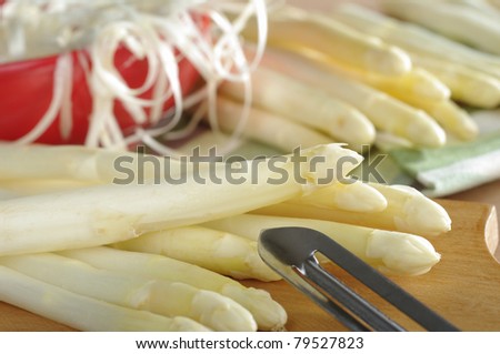 White asparagus with peeler on wooden board with asparagus and a red bowl full of peel in the back (Selective Focus, Focus on the asparagus lying on top)