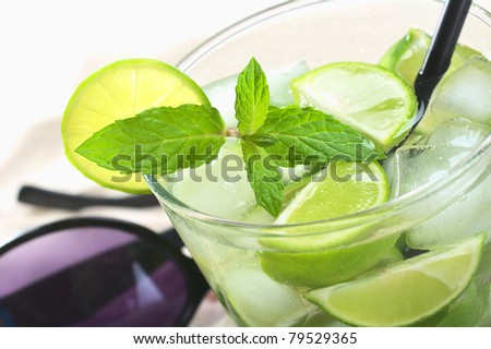 Mojito cocktail made of white rum, ice cubes, sugar, mint leaves and limes with a black drinking straw and sunglasses in the back (Selective Focus, Focus on the front of the mint leaf)
