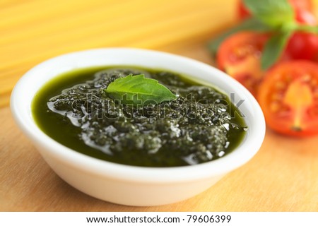 Fresh pesto made of basil, garlic and olive oil in a bowl garnished with basil leaf with raw spaghetti, cherry tomatoes and basil leaves in back (Selective Focus, Focus on the basil leaf in the bowl)