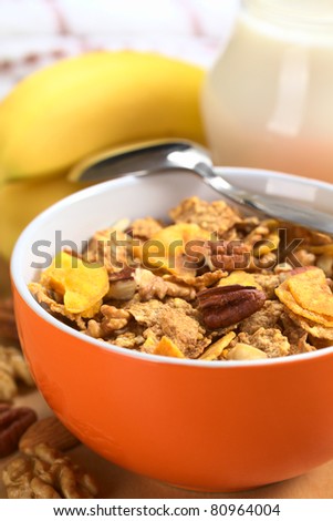 Breakfast with a bowl of wholewheat flakes with banana chips, walnuts and pecan nuts with nuts, banana and milk in a jug in the back (Selective Focus, Focus on the pecan nut one third into the bowl)