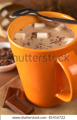 Hot chocolate with marshmallows in orange cup with a teaspoon on the rim and chocolate pieces on the side (Selective Focus, Focus on the marshmallows in the middle of the hot chocolate)