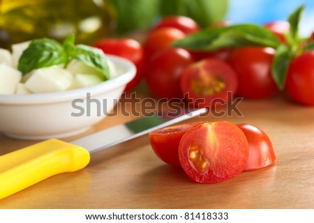 Raw cherry tomato cut in half with kitchen knife, cheese and basil in the back (Selective Focus, Focus on the surface of the cut tomato)