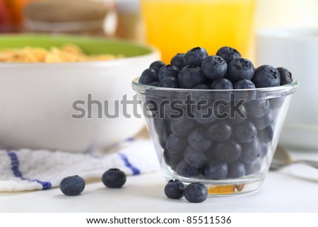 Fresh blueberries in glass bowl with a bowl of cereal, orange juice and cup in the back (Selective Focus, Focus on the blueberries in the front)