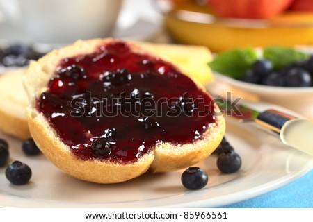 Blueberry jam on half a bun with fresh blueberries and other fruit and a cup in the back (Selective Focus, Focus on the left front of the jam and bun)
