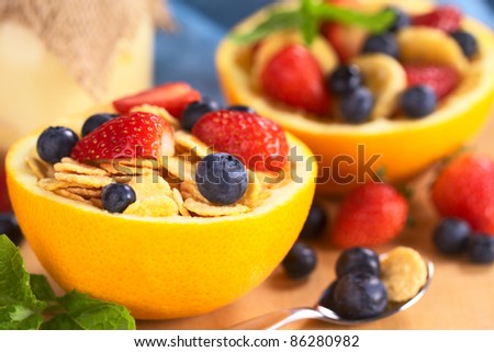 Corn flakes with fresh strawberries and blueberries served in a half orange bowl with yogurt and fruit salad in the back (Selective Focus, Focus on the blueberry on the right front of the cereal)