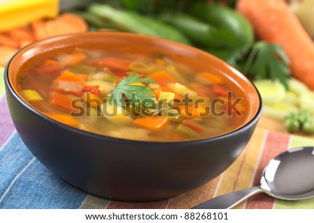 Vegetable soup made of green bean, pea, carrot, potato, red bell pepper, tomato and leek in black bowl garnished with a parsley leaf (Selective Focus, Focus on the front of the parsley leaf)