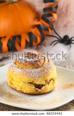 Pumpkin cinnamon roll garnished with confectioners\' sugar and cinnamon, Halloween decoration in the back (Selective Focus, Focus on the front edge of the roll)