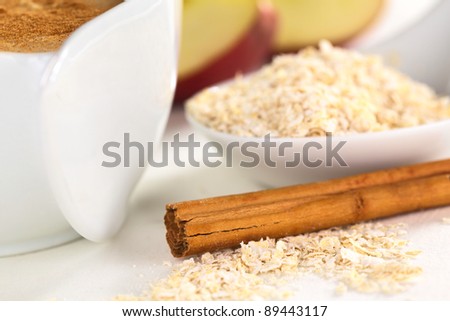 Cinnamon stick and oatmeal with porridge on the side and apple pieces in the back (Selective Focus, Focus on the front left end of the cinnamon stick and the oatmeal in the same plane)