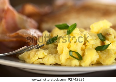 Scrambled eggs and green onions with fried bacon and toast bread in the back (Selective Focus, Focus on the green onion on the top and the egg in the same plane)