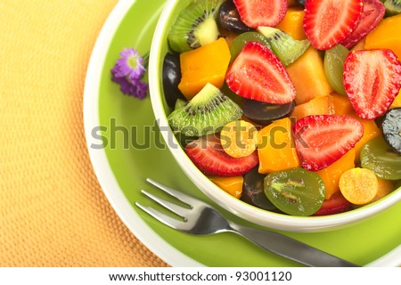 Fruit salad with strawberry, kiwi, grape, mango and physalis in a bowl with fork and small blue flowers photographed from above (Selective Focus, Focus from the front to the middle of the bowl)