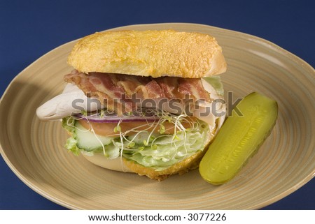 turkey club sandwich on a bagel,bacon,turkey,red onion,sprouts,tomato,cucumber,lettuce and a pickle, plate on blue background