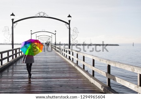 Woman holding a rainbow colored umbrella walking on a rainy day on the pier in White Rock, British Columbia, Canada.