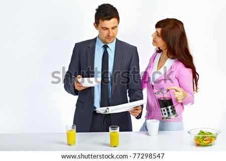 Business man reading newspaper in the kitchen before go to work and his wife holding kettle with coffee near him