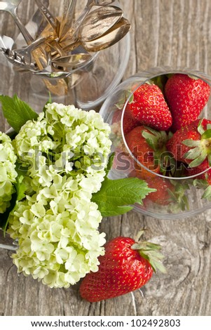 Top view on fresh strawberries, bunch of flowers and dessert tableware on old wooden table