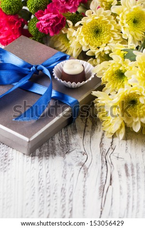 One chocolate candy on gray candy box with blue ribbon and yellow flowers on white wooden table