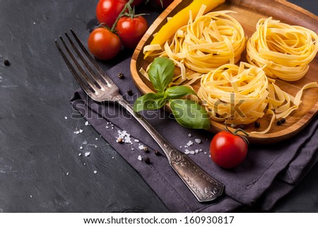 Wooden plate with dry pasta, fresh tomato, basil and yellow chili pepper served with sea salt and vintage fork on dark gray background