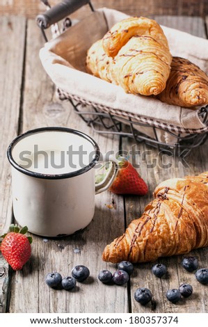 Fresh croissant with vintage mug of milk and berries over old wooden table.