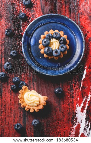 Top view on blueberry mini tarts served on blue ceramic plate over black and red wooden background.
